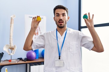 Young hispanic physiotherapist man holding strength balls to train hand muscles in shock face, looking skeptical and sarcastic, surprised with open mouth