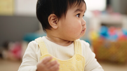 Adorable hispanic baby sitting on floor with relaxed expression at kindergarten