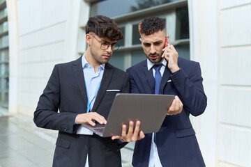 Two hispanic men business workers using laptop talking on smartphone at street