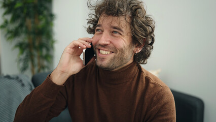 Young hispanic man talking on the smartphone sitting on sofa at home