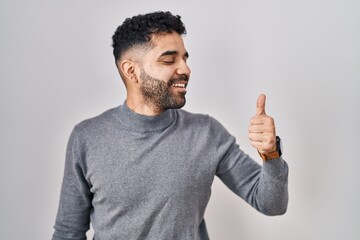 Hispanic man with beard standing over white background looking proud, smiling doing thumbs up gesture to the side