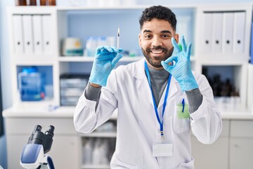 Hispanic man with beard working at scientist laboratory holding syringe doing ok sign with fingers, smiling friendly gesturing excellent symbol