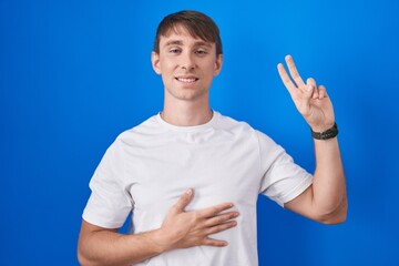 Caucasian blond man standing over blue background smiling swearing with hand on chest and fingers up, making a loyalty promise oath