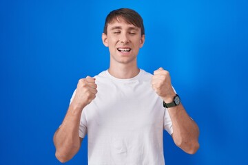 Caucasian blond man standing over blue background excited for success with arms raised and eyes closed celebrating victory smiling. winner concept.