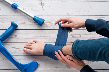 Teenage boy wrapping compression bandage around sprained ankle to reduce ache. Crutches on the...