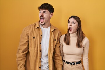 Young hispanic couple standing over yellow background angry and mad screaming frustrated and furious, shouting with anger. rage and aggressive concept.