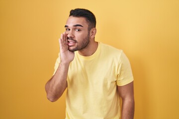 Young hispanic man standing over yellow background hand on mouth telling secret rumor, whispering malicious talk conversation