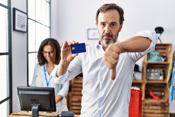 Hispanic man holding credit card at retail shop with angry face, negative sign showing dislike with thumbs down, rejection concept