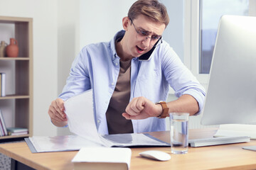 Emotional young man checking time while talking on phone in office. Being late