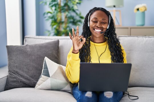 African woman working at home wearing operator headset doing ok sign with fingers, smiling friendly gesturing excellent symbol