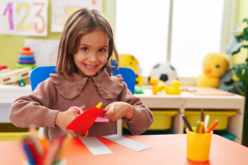 Adorable hispanic girl student smiling confident cutting paper at kindergarten