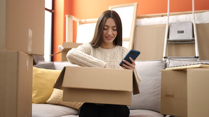 Young beautiful hispanic woman using smartphone unpacking cardboard box at new home