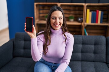Young brunette woman holding smartphone showing blank screen looking positive and happy standing and smiling with a confident smile showing teeth