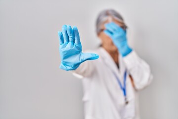 Middle age woman with grey hair wearing scientist robe covering eyes with hands and doing stop gesture with sad and fear expression. embarrassed and negative concept.