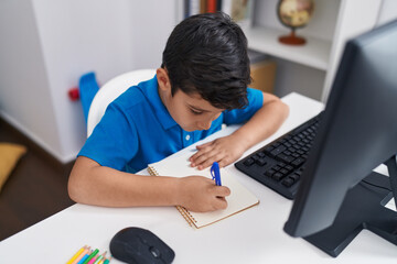 Adorable hispanic boy student using computer writing on notebook at classroom