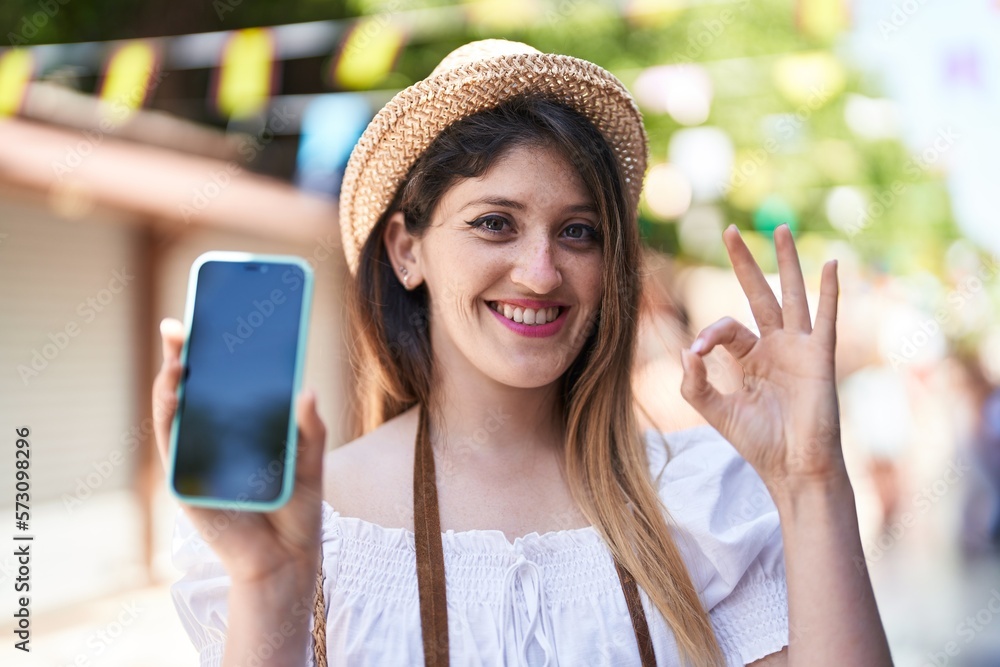 Wall mural Young brunette woman holding smartphone showing blank screen doing ok sign with fingers, smiling friendly gesturing excellent symbol