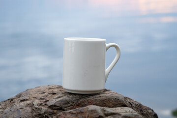 White mug, mock up, empty space for artwork, text, standing on stone, outdoors, green grass, trees, sky in the background, soft sunlight, nature