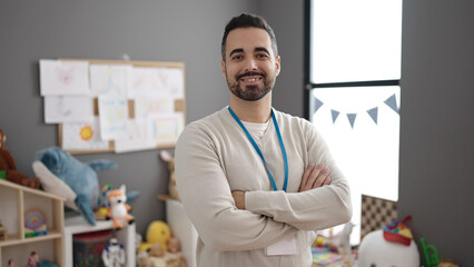 Young hispanic man teacher smiling confident standing with arms crossed gesture at kindergarten