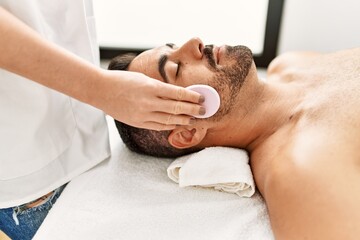 Obraz na płótnie Canvas Young hispanic man having clean facial treatment using sponge at beauty center