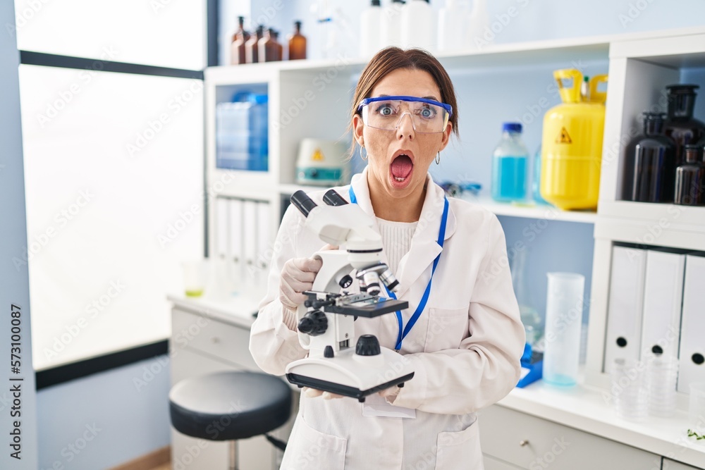 Sticker Young brunette woman working at scientist laboratory with microscope afraid and shocked with surprise and amazed expression, fear and excited face.