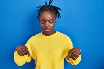 Beautiful black woman standing over blue background pointing down looking sad and upset, indicating...