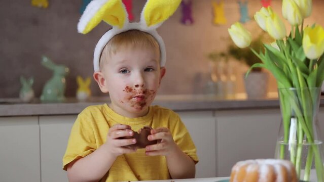 Little cute boy wearing bunny ears eating chocolate easter bunny while sitting at table at home.