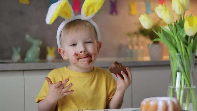 Little cute boy wearing bunny ears eating chocolate easter bunny while sitting at table at home.