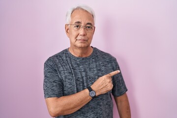 Middle age man with grey hair standing over pink background pointing with hand finger to the side showing advertisement, serious and calm face