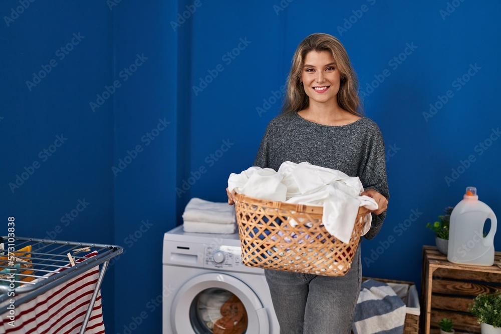 Wall mural young blonde woman smiling confident holding basket with clothes at laundry room