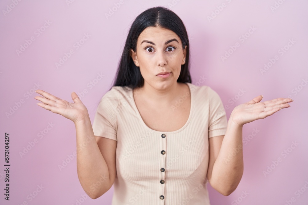 Poster Young hispanic woman standing over pink background clueless and confused expression with arms and hands raised. doubt concept.