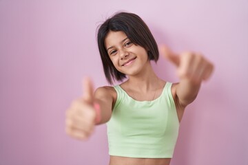 Young girl standing over pink background approving doing positive gesture with hand, thumbs up smiling and happy for success. winner gesture.