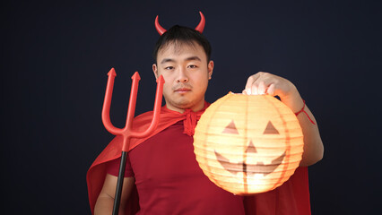 Young chinese man wearing devil costume holding pumpkin lamp over isolated black background