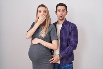 Young couple expecting a baby standing over white background shocked covering mouth with hands for mistake. secret concept.