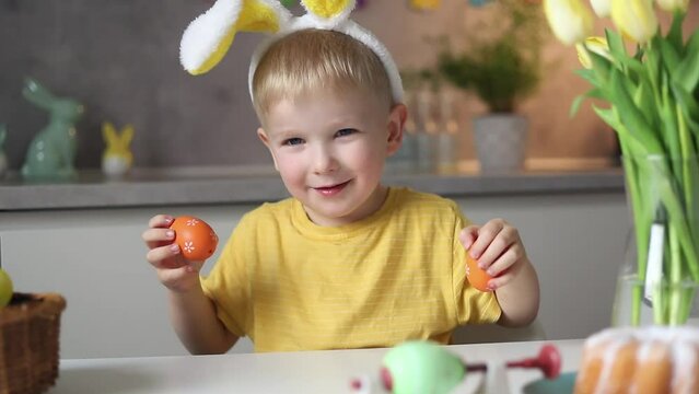 Emotional portrait of a cheerful little boy wearing bunny ears on Easter day who laughs merrily, plays with colorful Easter eggs sitting at a table in the kitchen.