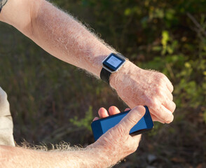 Senior man checking his heart rate and step count outdoor using his smart watch and smartphone; grass in the background