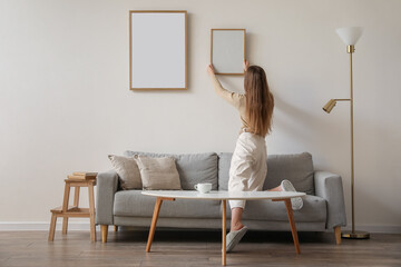 Young woman hanging blank frame on light wall at home, back view - Powered by Adobe