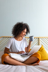 Fototapeta na wymiar Working at home. African American young woman sitting on bed drinking hot coffee while typing on laptop. Vertical