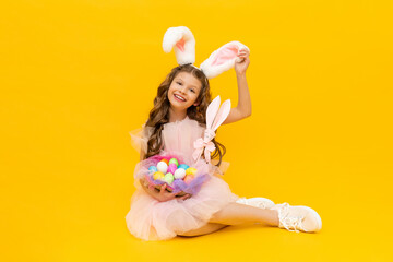 Festive Easter. A little girl with rabbit ears and a basket of colorful eggs smiles broadly on a yellow isolated background.