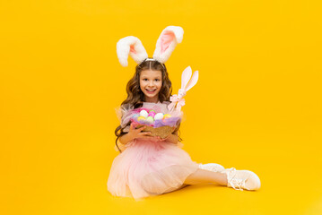 Festive Easter. A little girl with rabbit ears and a basket of colorful eggs smiles broadly on a yellow isolated background.