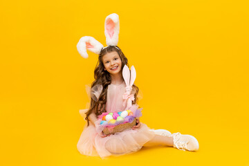 Festive Easter. A little girl with rabbit ears and a basket of colorful eggs smiles broadly on a yellow isolated background.