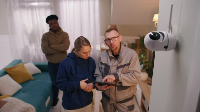 Installer sets up security camera at home of multi ethnic family using digital tablet. Man checks motion sensor of CCTV camera in professional app. Concept of tracking system, safety and privacy.