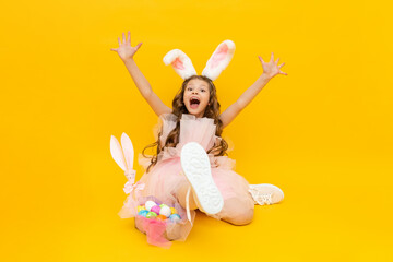 Festive Easter. A little girl depicts an Easter bunny with a basket of colorful eggs. A very happy child with rabbit ears on a yellow isolated background.