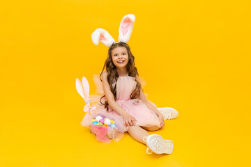 A little girl dressed as an Easter bunny with a basket of colorful eggs on a yellow isolated background. Festive spring Easter.