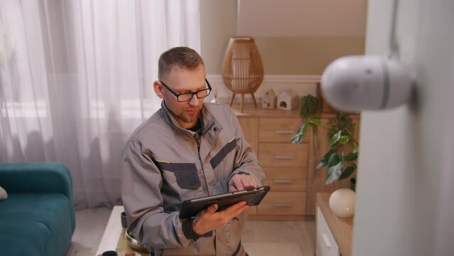 Male installer in uniform sets up security camera in apartment using digital tablet computer. Man checks motion sensor of CCTV camera in application. Concept of tracking system, safety and privacy.