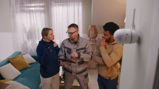 Installer sets up security camera in apartment using digital tablet. Man shows to multi cultural couple how to check motion sensor of CCTV camera in app. Tracking system, safety and privacy concept.