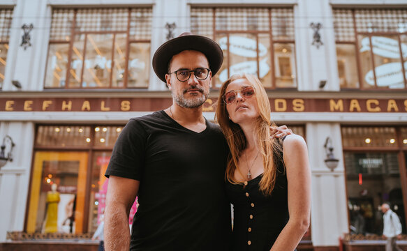 Handsome young beardy man in glasses, hat , black clothes embracing redhead girlfriend in city, European couple in vacation trip walking, looking at camera. Confident carefree people. Journey, trip.