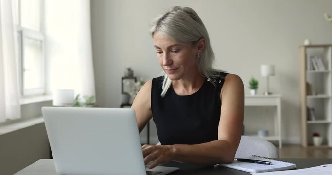 Focused mature woman working or studying on laptop, gains new knowledge, improving professional skills, search helpful information on internet, jotting in copybook sit at desk in home office. E-learn