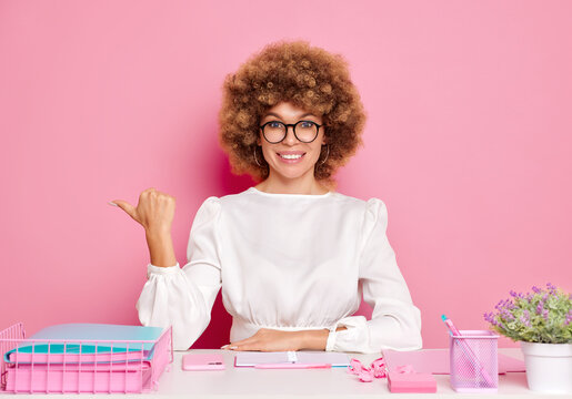 Positive woman employee points thumb finger aside to copy space, looks in the camera, has new idea, dressed in bright formal blouse, sits at desktop in office, isolated over pink wall. People and job