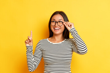 Indoor shot of amazed European brunette woman in transparent eyewear stares with happy expression points index finger up shows some promotion wears striped t-shirt poses against yellow wall. People