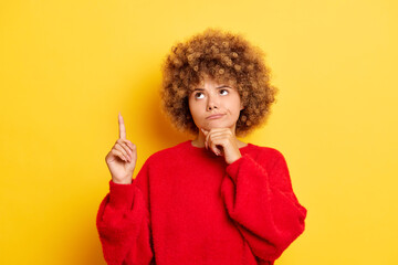 Horizontal shot of cute thoughtful young woman with curly hair points index finger up demonstrates something wears red pullover poses over yellow background. Promotional concept